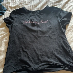 Svart t-shirt Victoria's Secret M - Svart t-shirt från Victoria's Secret i mjuk bomull. Klassisk passform med korta ärmar och rund hals. Framsidan har en snygg rosa logga i skrivstil. Perfekt för en chill och stilren look.