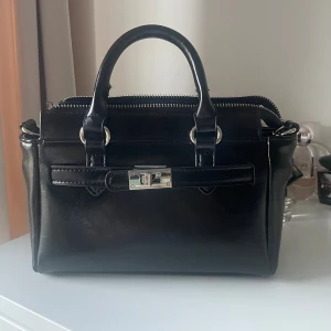 Svart handväska  - Stilren svart handväska ifrån Zalando i skinn med två handtag, silverfärgad dragkedja och snyggt spänne framtill. Väskan har flera fack inuti, inklusive ett med dragkedja, perfekt för att hålla ordning på dina grejer. Klassisk och tidlös design som passar till det mesta.