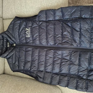 Svart dunväst från EA7 Emporio Armani - Snygg svart dunväst från EA7 Emporio Armani med quiltad design och dragkedja framtill. Västen har hög krage och logga på bröstet. Västen har ett mycket bra skick använd ett fåtal gånger.
