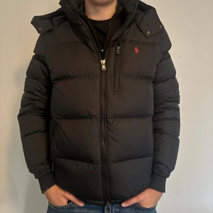 Svart pufferjacka Polo Ralph Lauren M - Svart pufferjacka från Polo Ralph Lauren med röd logga på bröstet. Endast använd under förra vintern och är i toppskick. Enda defekten är ett ca 0.5 cm stort hål som numera är lagat. Det sitter på undersidan av högra armen och märks knappt av. Finns det några övriga funderingar så är det bara att höra av sig! Vid snabb affär kan pris diskuteras