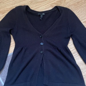 Svart kofta med v-ringning från H&M - Svart stickad kofta från H&M i storlek XS. Koftan har v-ringning, tre knappar framtill och långa ärmar med ribbade detaljer. Perfekt att slänga över en topp för en chill look. Enkel och stilren design som funkar året runt.