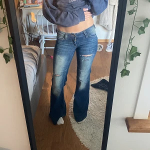 Blå bootcut jeans från Cheap Monday - Snygga blå bootcut jeans från Cheap Monday med slitningar och hål på låren. Låg midja och klassisk femficksmodell. Jeansen har en cool vit patch med döskalle på bakfickan och orange detalj vid linningen. Perfekta för dig som gillar en avslappnad och trendig look.