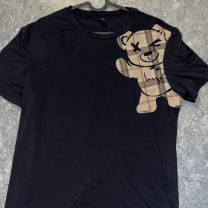 Svart t-shirt med nalletryck - Svart t-shirt med ett stort tryck av en rutmönstrad nalle på axeln. T-shirten har rund hals och korta ärmar. Materialet känns mjukt och skönt, troligtvis bomull eller bomullsblandning. Perfekt för dig som gillar streetstyle och unika prints.