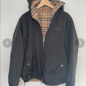 Burberry vindjacka med huva, L - Burberry vindjacka i svart med klassiskt beige rutigt foder. Jackan har huva, dragkedja framtill, snörning i nederkant och tryckknappar vid fickorna. Utsidan är svart och insidan har ikoniskt Burberry-mönster. Tillverkad i bomull.