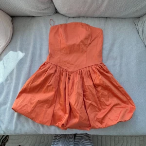 Axelbandslös orange kortklänning - Säljer en axelbandslös orange kortklänning med voluminös kjol och markerad midja. Klänningen har en snygg, enkel design och är tillverkad i ett lätt tyg som ger en luftig känsla. Liten i storleken 