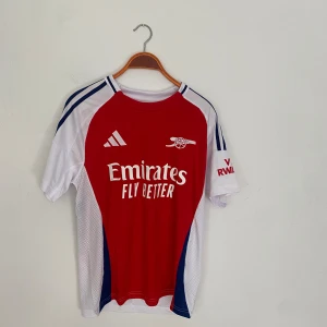 Arsenal Adidas röd vit fotbollströja - Snygg Arsenal fotbollströja från Adidas med klassisk röd front och vita ärmar. Tröjan har blå detaljer och tre ränder på axlarna, samt klubbens logga och Emirates-sponsorn tryckt på bröstet. Tillverkad i lätt och ventilerande material.