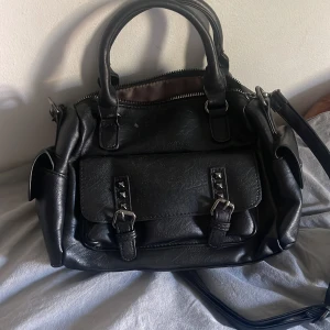 Svart axelväska med spännen - Säljer en svart axelväska i skinnimitation med både handtag och lång axelrem. Väskan har flera fack, dragkedja upptill och snygga detaljer med spännen och nitar på framsidan. Perfekt storlek för att få plats med allt du behöver till skolan eller stan.PRIS KAN DISKUTERAS 