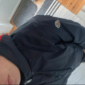 Mörkblå vindjacka från Moncler - Säljer en mörkblå vindjacka från Moncler med huva och snygga röda och vita detaljer på luvan. Jackan har dragkedja framtill, två fickor med knappar och Moncler-logga på ärmen. Perfekt för blåsiga dagar och enkel att matcha med det mesta.