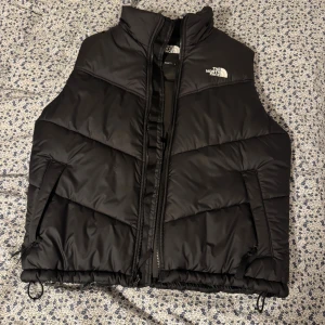  The North Face-Saikuru Väst. HELT NY, Nypris 2200kr - Säljer en svart dunväst från The North Face med hög krage och dragkedja framtill. Västen har två snedställda fickor med dragkedja och justerbar dragsko nedtill. Klassisk logga på bröstet och ryggen. Perfekt för lager-på-lager under kyliga dagar. Nypris:2200kr, Endast använd 1 gång