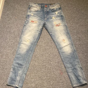Jack & Jones Erik Anti Fit jeans 30/32 - Ljusblå slitna jeans från Jack & Jones, modell Erik Anti Fit. Jeansen har rött tyg bakom slitningarna och har Slim straight passform, flera slitningar och lagningar framtill samt klassiska fem fickor. Materialet är denim i bomull och de har en cool, avslappnad vibe.