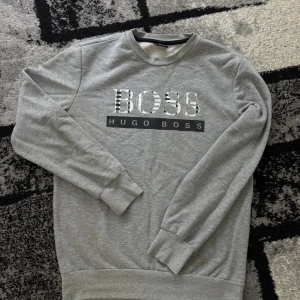 Boss hugo boss tröja - Grå sweatshirt från Hugo Boss med stort BOSS-tryck i svartvitt mönster på bröstet och klassisk rund hals. Tröjan har långa ärmar, ribbade muddar och är tillverkad i mjuk bomullsmix. Perfekt för en clean och stilren look.