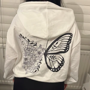 Vit hoodie med fjäril och blommor - Vit hoodie med stor svart fjärilsvinge och blommigt mönster på ryggen. Tröjan har huva med snörning och långa ärmar. Oversized passform som ger en chill vibe. Perfekt för dig som gillar unika tryck och vill sticka ut lite extra.