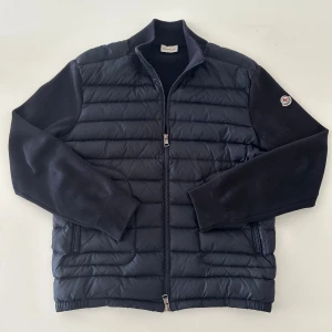 Moncler cardigan  - Säljer en marinblå cardigan från Moncler med stickade ärmar och quiltad front. Jackan har dragkedja, ribbade muddar och Moncler-logga på ena ärmen. Fylld med vitt gåsdun för extra värme. Perfekt för dig som vill ha en clean och stilren look. Hör av er vid frågor