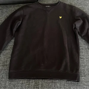 Svart sweatshirt från Lyle & Scott - Svart sweatshirt från Lyle & Scott Junior med klassisk rund hals och gul broderad logga på bröstet. Tröjan har långärmad passform och är tillverkad i mjuk bomull, perfekt för en chill och stilren look.