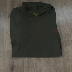 Olivgrön t-shirt från Ralph Lauren - Snygg olivgrön t-shirt från Ralph Lauren med klassisk röd broderad logga på bröstet. Modellen är kortärmad och har en rund hals. Tillverkad i mjuk bomull som känns skön mot huden. Perfekt för dig som gillar stilrena och enkla plagg med en touch av lyx. Tröjan är i nyskick!