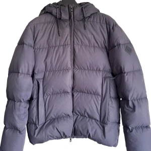 Blå pufferjacka från Gant med huva - Säljer en blå pufferjacka från Gant, modell Alta Down Jacket. Jackan har huva, dragkedja framtill och stora fickor. Fylld med dun och fjädrar, vattenavvisande och andas. Perfekt för kalla vinterdagar och riktigt skön att bära.