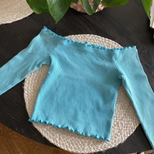 Turkos offshoulder topp - Gina Tricot - Superfin turkos ribbad offshoulder topp från Gina Tricot i storlek S. Långärmad modell med volangkant vid halsringning, ärmslut och nederkant. Toppens material är stretchigt.