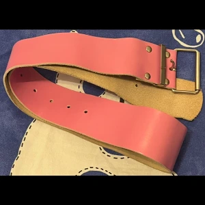 Brett rosa skärp i skinn - Säljer ett brett rosa bälte i skinn med stilren, fyrkantig metallspänne och nitar. Bältet har en slät yta och flera hål för justering. Perfekt för att ge outfiten en färgklick och cool vibe. Cirka 105 cm långt.