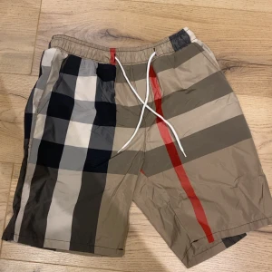 Burberry simshorts - Snygga shorts med klassiskt rutigt mönster i beige, svart, vitt och rött. De har elastisk midja med vit snörning för skön passform. Perfekta för sommaren och har en riktigt trendig vibe. Materialet känns lätt och luftigt, perfekt när det är varmt ute.