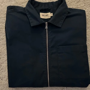Svart overshirt från East West - Snygg svart overshirt från East West med dragkedja framtill och en praktisk bröstficka. Perfekt för lager-på-lager-stil och passar till många olika outfits.