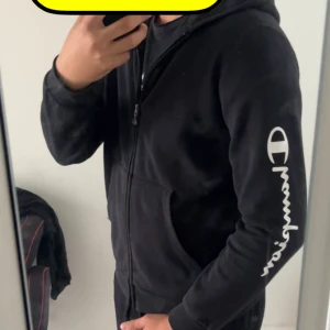 Svart Champion hoodie med dragkedja - Svart hoodie från Champion i storlek XL med vit logga på bröstet och ärmen. Tröjan har huva, dragkedja framtill och två fickor. Tillverkad i mjuk bomullsmix, perfekt för chill eller streetwear. Klassisk och enkel design som funkar året runt.