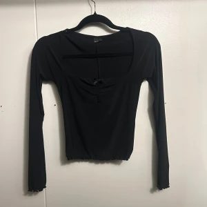 Svart långärmad topp Gina Tricot S - Svart långärmad topp från Gina Tricot i stretchigt material. Modellen har en söt liten rosett framtill, rynkade detaljer vid bysten och lätt vågiga kanter vid ärmslut och nederkant. Djup, rundad ringning och figurnära passform. Perfekt till jeans eller kjol.