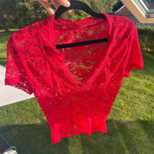 Röd spetsblus med v-ringning - Säljer en röd, transparent spetsblus med blommigt mönster och djup v-ringning. Blusen har korta ärmar och är figurnära i modellen. Perfekt för dig som vill sticka ut med en färgstark och romantisk look.