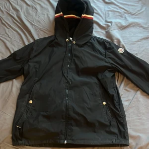 Svart vindjacka från Moncler med huva - Svart vindjacka från Moncler med huva och coola rödvita ränder på huvan. Jackan har dragkedja framtill, två fickor med silverknappar och Moncler-logga på ärmen. Materialet är lätt och syntetiskt, perfekt för blåsiga dagar. Snygg och sportig look. Finns dock lite brister då ena saken på dragkedjan är av och ett litet hål vid vänster ficka