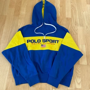 Blå och gul hoodie Polo Sport - Snygg blå hoodie från Polo Sport Ralph Lauren med breda gula paneler över bröst, rygg och ärmar. Stor logga framtill och amerikansk flagga. Tröjan har huva med vit snörning och är tillverkad i bomull och polyester. Perfekt för dig som gillar sportig och färgstark stil.