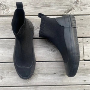 Svarta boots från Tommy Hilfiger - Snygga svarta boots från Tommy Hilfiger med stickad ovandel och robust gummisula. Skorna har rund tå, dragflik baktill med Hilfiger-logga och diskret flagga på hälen. Perfekta för dig som gillar en clean och modern look.