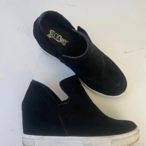 Svarta wedge sneakers från Trash - Svarta sneakers med kilklack från Trash.Skorna har en rund tå, platt vit sula och är tillverkade i mocka. Perfekta för dig som vill ha lite extra höjd men ändå en sportig look.