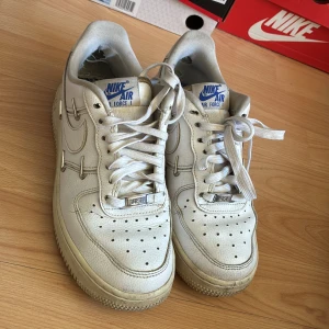 Nike Air Force 1 vita sneakers - Klassiska Nike Air Force 1 sneakers i vitt skinn med blå logga på plösen och coolt mönstrad innersula. Lågt skaft, perforerad tå och snörning. Snygga detaljer med metall på snörningen och robust sula. Perfekta för dig som gillar streetstyle.