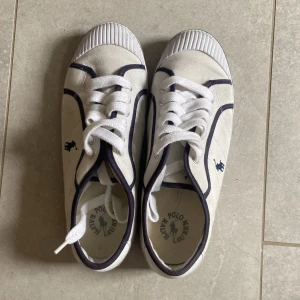 Vita sneakers från Polo Ralph Lauren storlek 39 - Snygga vita sneakers från Polo Ralph Lauren med mörkblå detaljer och broderad logga på sidan. Skorna har klassisk snörning, rund tå och platt sula. Tillverkade i bomullscanvas med gummidetaljer på tån och sulan. Perfekta för en clean och sportig stil. PRISET GÅR ATT DISKUTERAS!