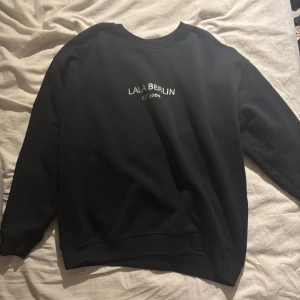 Svart sweatshirt från Lala Berlin - Svart sweatshirt från Lala Berlin med vit text 'LALA BERLIN EST 2004' på bröstet. Klassisk rund halsringning och långa ärmar. Mjuk insida och relaxed fit, perfekt för en chill och stilren look.