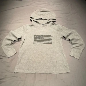 Grå hoodie från Polo Ralph Lauren - Grå hoodie från Polo Ralph Lauren med svart flaggtryck och text på bröstet. Tröjan har huva, långa ärmar och är tillverkad i mjuk bomullsmix. Perfekt för en avslappnad och stilren look.