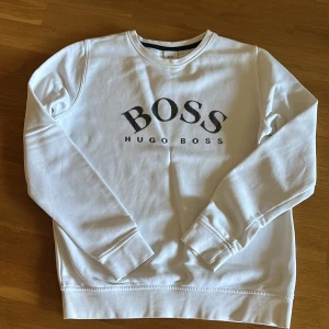 Vit sweatshirt från Hugo Boss - Vit sweatshirt från Hugo Boss med stor svart logga på bröstet. Tröjan har rund halsringning och långa ärmar. Tillverkad i mjuk bomullsblandning med lite polyester för extra komfort. Perfekt för en clean och stilren look.