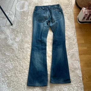 Blå bootcut jeans från G-Star - Snygga blå jeans från G-Star med bootcut passform och klassisk femficksdesign. Jeansen har en lätt tvättad look och coola detaljer på bakfickorna. Perfekta för dig som gillar en avslappnad men trendig stil. Ett hål vid innerbenet men som enkelt går att sy igen.