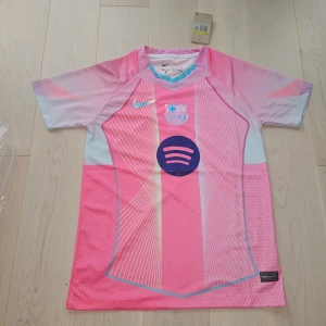 Barça x Nike rosa fotbollströja - Säljer en FC Barcelona fotbollströja från Nike i rosa och ljusblått med vita och gula linjer. Tröjan har korta ärmar, V-ringning och tryckta loggor från både Nike och Barça samt Spotify som sponsor. Tillverkad i lätt och ventilerande Dri-FIT-material.