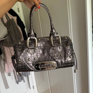 Guess metallic handväska med crocomönster - Säljer en metallic-silver handväska från Guess med crocomönstrad yta och coola nitar. Väskan har två rejäla handtag, dragkedja upptill och snygga metalldetaljer. Insidan är fodrad med mönstrat tyg i brunt och lila. Perfekt för dig som vill sticka ut.