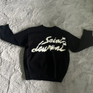 Svart Saint Laurent stickad tröja - Svart stickad tröja från Saint Laurent med vit, fuzzy logga framtill. Tröjan har rund halsringning och långa ärmar. Perfekt för dig som gillar streetstyle och vill sticka ut med en ikonisk design.