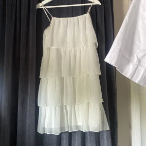 Vit volangklänning från Vila - Superfin vit kortklänning från Vila med tunna axelband och flera lager volanger. Klänningen är luftig och har en flowy känsla, perfekt för varma dagar. Tillverkad i 100% polyester och har en lätt, transparent look.