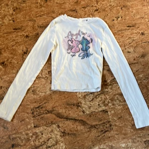 Vit långärmad ribbad tröja H&M - Vit ribbad långärmad tröja från H&M med tryck framtill av två tecknade figurer och texten 'Angel' och 'Silly' i rosa och blått. Tröjan har en croppad passform och är tillverkad i mjuk bomull. Perfekt för dig som gillar gulliga motiv och bekväma basplagg.