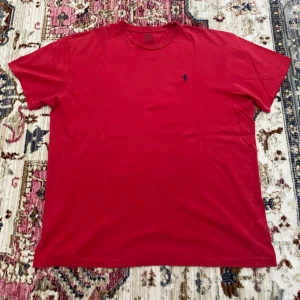 Röd t-shirt från Polo Ralph Lauren - Röd t-shirt från Polo Ralph Lauren med klassisk rund hals och blå broderad logga på bröstet. T-shirten är i bomull och har en enkel, stilren design med korta ärmar. Perfekt för en clean och avslappnad look.