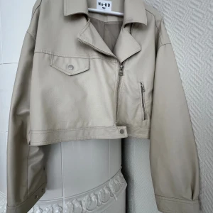 Beige croppad skinnjacka från NA-KD - Snygg beige croppad PU jacka från NA-KD med klassisk krage, dragkedja och coola detaljer som fickor med lock och knapp. Jackan har långa ärmar och en loose fit som ger en chill vibe. Perfekt för dig som vill ha en trendig look.