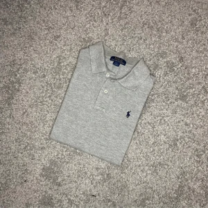 Ralph Lauren långärmad piké  - Stilren grå långärmad piké från Ralph Lauren – en perfekt kombination av sportig elegans och komfort. Tillverkad i mjuk bomull med klassisk krage, knappar och den ikoniska broderade loggan på bröstet. Ett mångsidigt plagg som passar året runt – lika snygg till jeans som chinos. Inga fläckar eller slitage – fräsch och redo för användning!