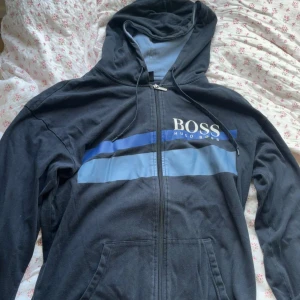 Mörkblå  hoodie från Hugo Boss med blå detaljer - Svart hoodie från Hugo Boss med dragkedja och huva. Framsidan har blå och ljusblå ränder samt stor BOSS-logga. Tröjan har snörning i huvan och fickor framtill. 