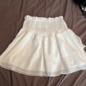 Vit volangkjol  - Supersöt vit kortkjol med bred smock i midjan och dubbla volanger. Kjolen har ett lätt och flowigt tyg som ger en luftig känsla. Har aldrig använt tyvärr men jätte fin