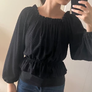 Svart blus med volang och struktur - Svart blus med volangkant i midjan och vid halsringningen. Toppens tyg har små prickar i strukturen och vida, långa ärmar med resår vid handlederna. Sitter snyggt med smock i midjan och ger en romantisk vibe. Perfekt att styla med jeans eller kjol.