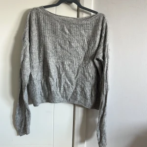 Grå stickad tröja med bred hals - Mysig grå stickad tröja med bred halsringning och lång ärm. Tröjan har en avslappnad passform och ribbade muddar vid ärmslut och nederkant. Perfekt för lager-på-lager och chill dagar.