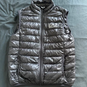 Svart dunväst från Emporio Armani EA7 - Snygg svart dunväst från Emporio Armani EA7 med glansig finish och horisontella quiltade sömmar. Västen har hög krage, dragkedja framtill och logga på bröstet. Perfekt för lager-på-lager och streetstyle.